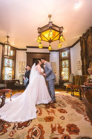 Bride and groom share first kiss in elegant Victorian room with stained glass windows and chandelier.