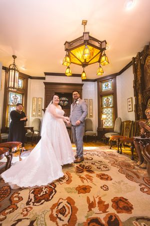 Bride and groom exchange vows in ornate Victorian parlor with stained glass windows and chandelier.