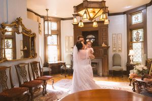 Bride and groom share a romantic embrace in an elegant Victorian parlor with ornate gold mirrors and stained glass windows.
