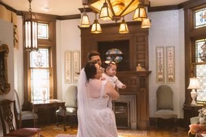 Bride and groom hold their baby in an elegant Victorian parlor with stained glass windows and chandelier.