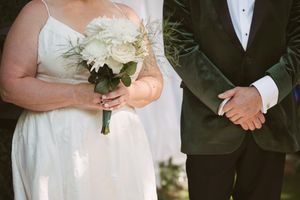 Bride holding white rose bouquet with greenery beside groom in dark green velvet tuxedo at outdoor wedding ceremony.