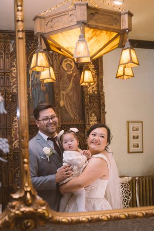 Smiling newlyweds in wedding attire hold their baby girl, reflected in an ornate gold mirror with warm pendant lights.