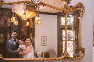Couple embraces with child reflected in an ornate gold-framed mirror at a vintage wedding venue.