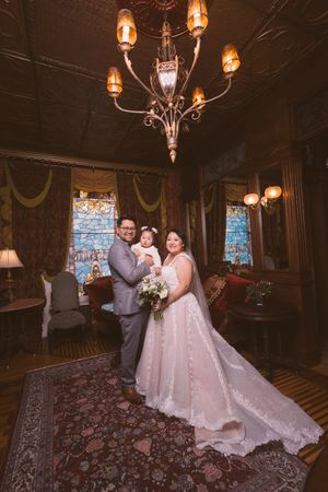 Bride and groom holding baby in elegant Victorian room with chandelier and stained glass windows on wedding day.