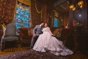 Bride and groom share romantic moment on ornate sofa in elegant Victorian room with stained glass windows.