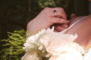Close-up of bride's hands with emerald engagement ring holding white rose wedding bouquet outdoors.