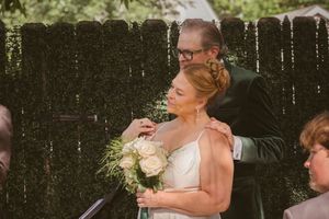 Groom in dark suit embracing bride holding white bouquet during outdoor wedding ceremony.