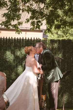Bride and groom share their first kiss outdoors beneath green trees in front of a lush hedge wall.