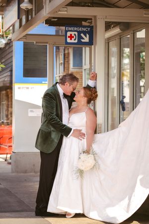 Bride and groom share romantic kiss outside hospital emergency entrance, brides veil flowing in the breeze.
