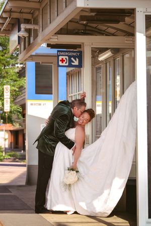 Groom dips and kisses bride outside a transit station entrance beneath an emergency sign on wedding day.