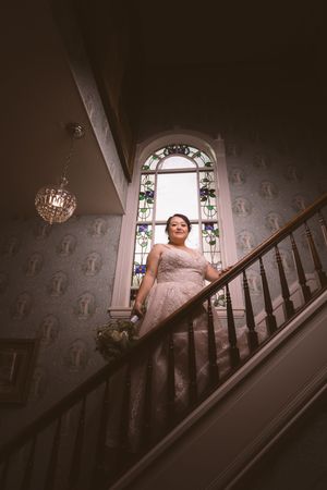 Bride in lace gown poses on grand staircase beneath arched stained glass window in historic venue.