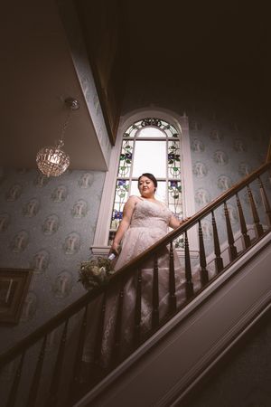 Bride in lace gown descends grand staircase beneath stunning arched stained glass window in historic mansion.