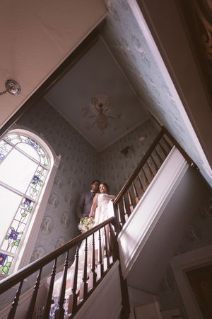 Bride and groom stand on elegant staircase with stained glass window in historic wedding venue.