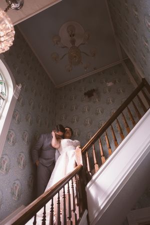 Bride and groom share a romantic kiss on an elegant staircase with vintage patterned wallpaper and chandelier.