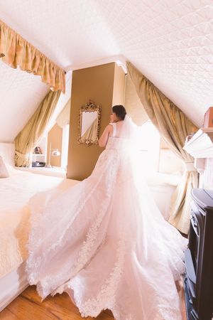 Bride in elegant lace wedding gown with long train standing in sunlit bridal suite before ceremony.