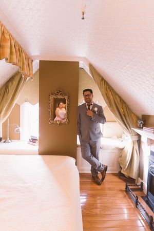Groom in gray suit adjusting boutonniere in elegant attic bedroom with hardwood floors and ornate mirror.