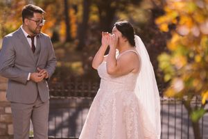 Emotional bride covers her face with hands during outdoor first look with groom amid autumn foliage.