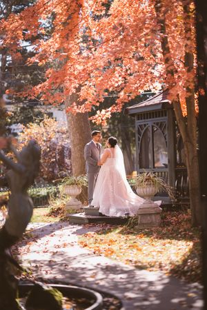 Bride and groom share a romantic moment under vibrant autumn foliage in a garden with a gazebo.