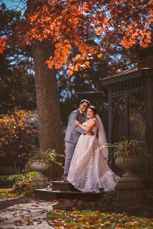 Bride and groom embrace under vibrant autumn maple tree beside gazebo in romantic fall wedding portrait.