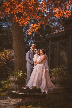 A couple in wedding attire holds their baby under vibrant fall foliage beside a gazebo.