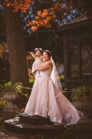 Bride in lace gown holds baby girl in matching dress amid autumn foliage at outdoor wedding venue.