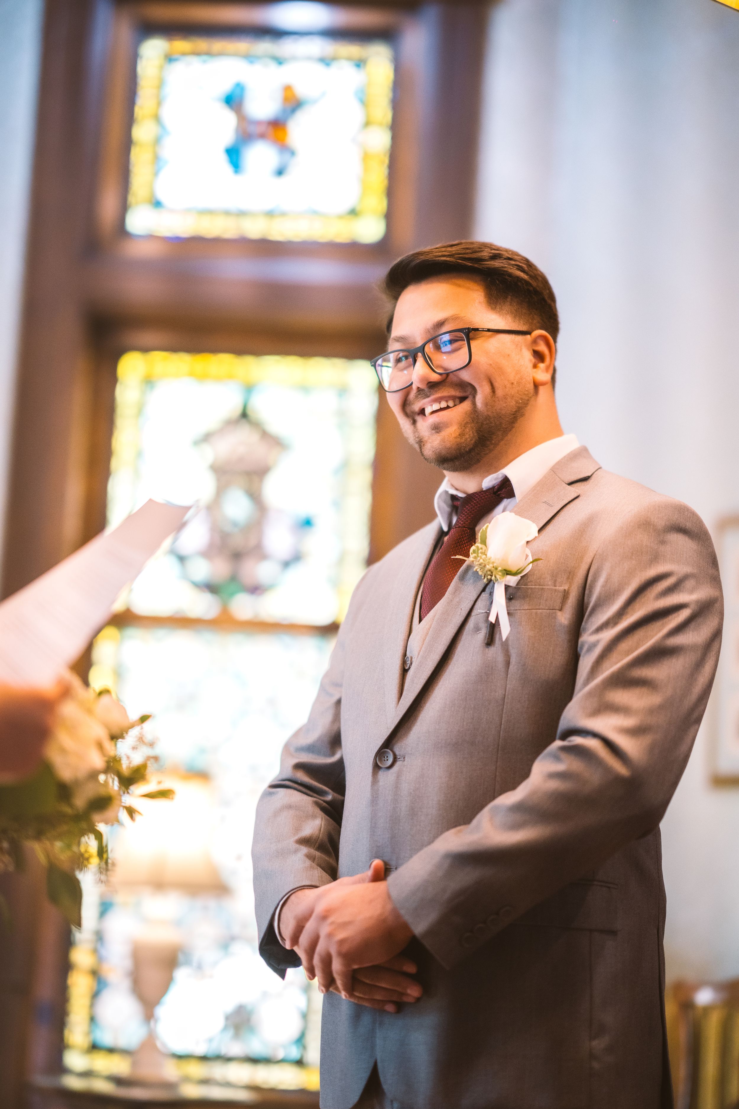 Smiling groom in gray suit with boutonniere stands at altar before stained glass windows during ceremony.