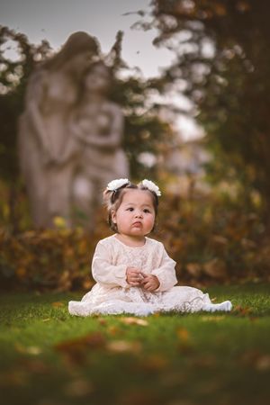 Adorable baby girl in white lace dress sits on green grass with angel statue in soft background.