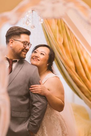 Smiling bride in lace gown embraces groom in gray suit with glasses under elegant golden drapery.