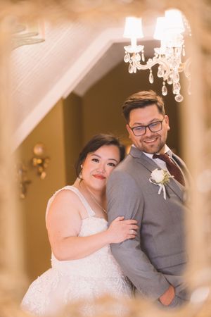 Smiling bride and groom pose together in mirror reflection beneath a crystal chandelier on their wedding day.