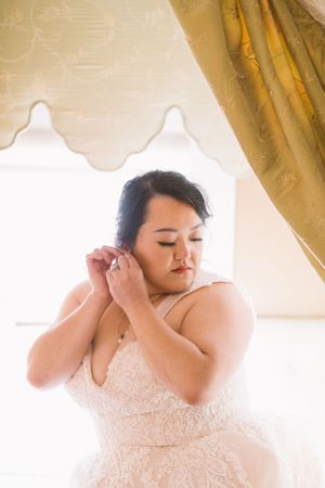 Bride in lace wedding gown adjusts earring near sunlit window with elegant golden drape backdrop.