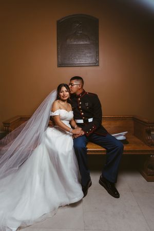 Military groom in Marine dress uniform sits with bride in white wedding gown and veil on wooden bench.