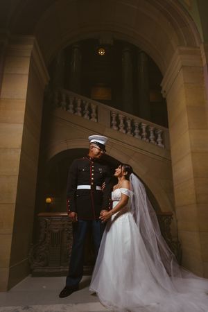 Marine groom in dress uniform stands with bride in white gown and veil inside elegant stone building.