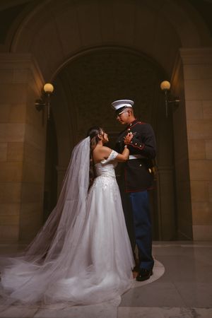 Military groom in Marine dress blues holds bride in white wedding gown under stone archway in romantic pose.