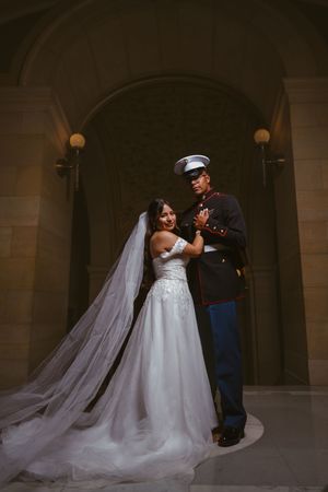 Bride in lace wedding gown with flowing veil embraces Marine groom in dress blues under stone archway.