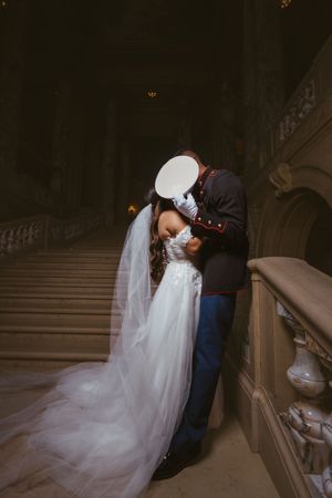 Marine groom in dress uniform kisses bride in white gown on grand staircase in romantic wedding photo.