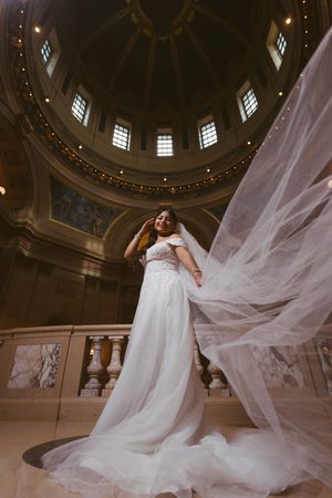 Bride in white gown poses under grand dome with veil flowing dramatically in historic building.