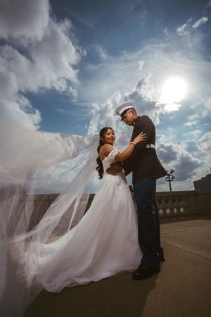 Marine groom holds bride in white gown as her veil billows in wind against dramatic cloudy sky.