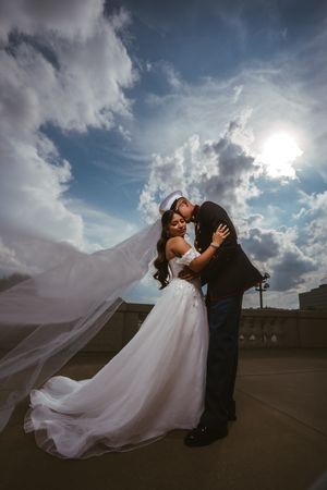 Bride and groom kiss outdoors with wedding veil flowing in wind against dramatic blue sky.