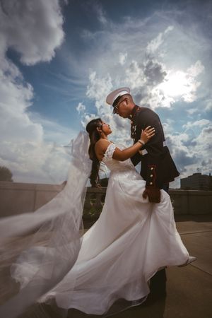 Marine groom in dress uniform dances with bride outdoors under dramatic cloudy blue sky.