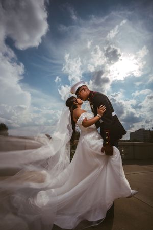 Marine groom kisses bride in flowing white gown outdoors as her veil sweeps dramatically in the wind.