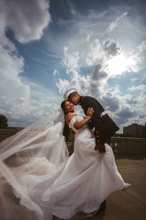 Marine groom in dress uniform kissing bride in flowing white gown on rooftop under dramatic cloudy sky.