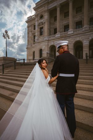 A bride in a flowing white gown and veil holds hands with a Marine in dress uniform on capitol building steps.