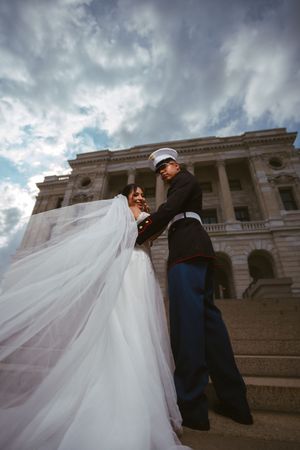 Marine groom holds bride on steps of neoclassical building as her veil flows in the wind.