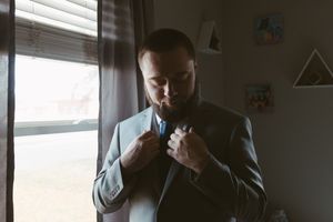 A bearded groom in a gray suit adjusts his blue tie while standing near a window with soft natural light.