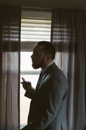 A bearded man in a suit stands by a window with curtains, holding a phone in dramatic side lighting.