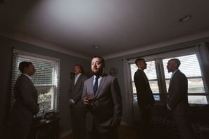 Groomsmen in suits gather in a softly lit room, with one man standing in the foreground holding a drink.