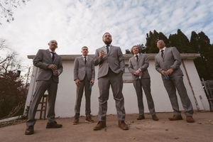 Five groomsmen in matching gray suits pose outdoors against a white wall under a cloudy sky.
