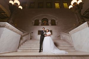 Bride and groom pose elegantly on grand marble staircase in ornate wedding venue with warm chandelier lighting.
