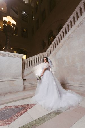 Bride in a flowing white gown and veil poses gracefully on marble stairs in an elegant grand hall.
