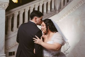 Bride and groom share an intimate moment on grand staircase, bride in white lace gown with veil, groom in black tuxedo.